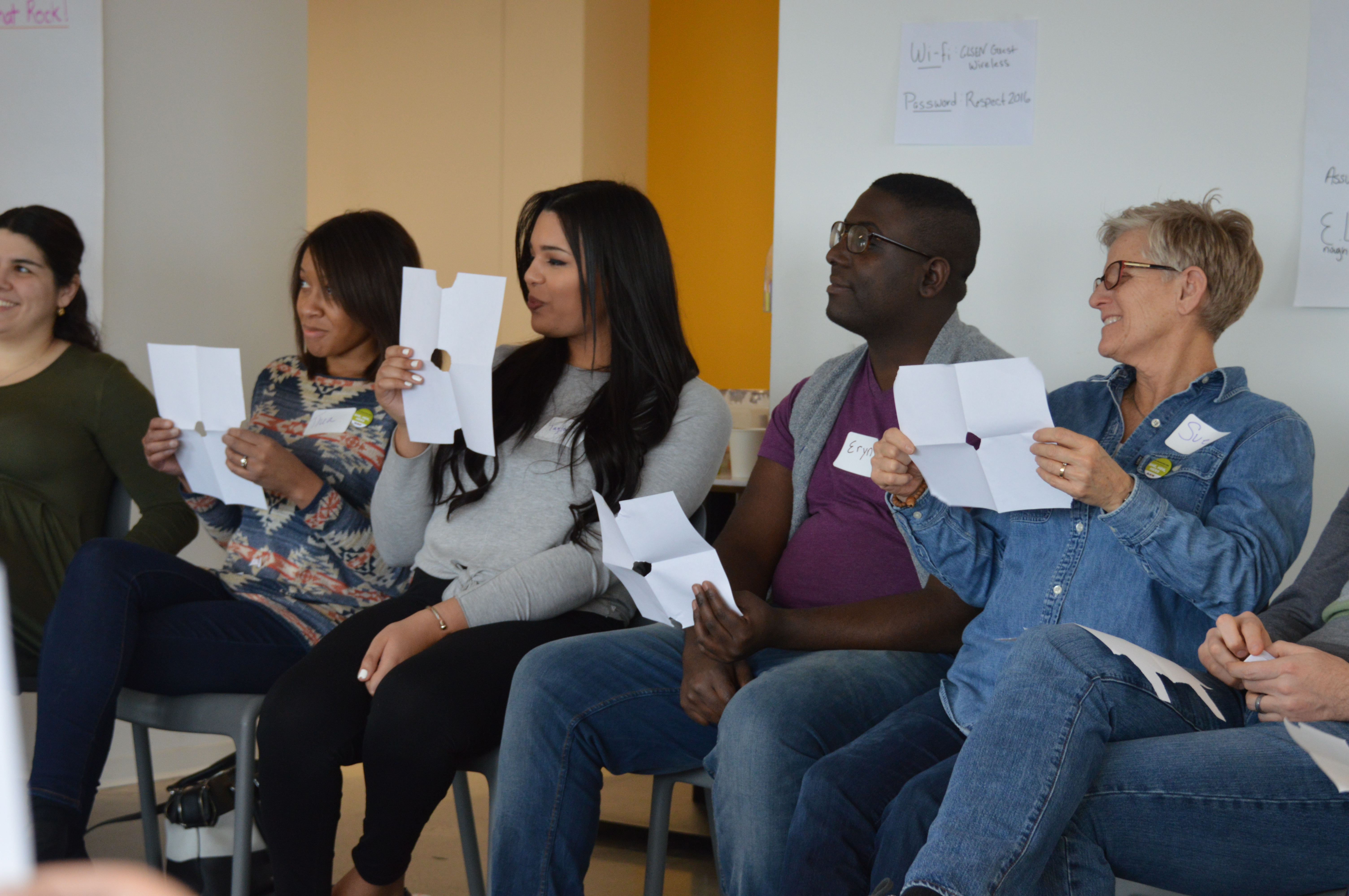 Four sitting adult volunteers participate in an activity at GLSEN's NYC Training of Trainers, or TOT. They smile and hold up pieces of white paper with fold lines and a circle cut in the center.