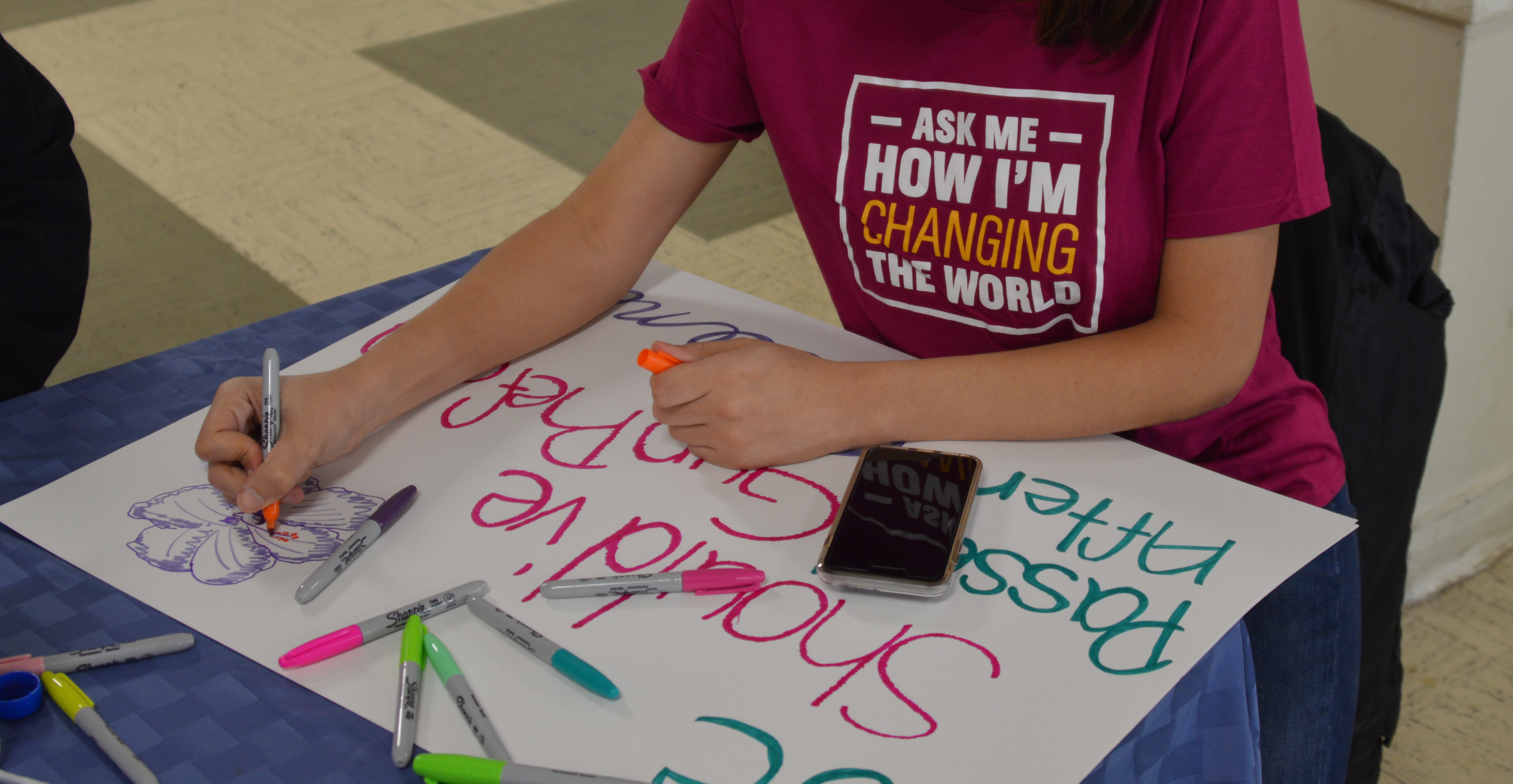 A GLSEN volunteer sits at a table and works on their protest sign for the Washington, D.C. March for Our Lives demonstration. They wear a GLSEN t-shirt that reads: Ask me how I'm changing the world. On the table are a cell phone and several Sharpie markers in various colors.