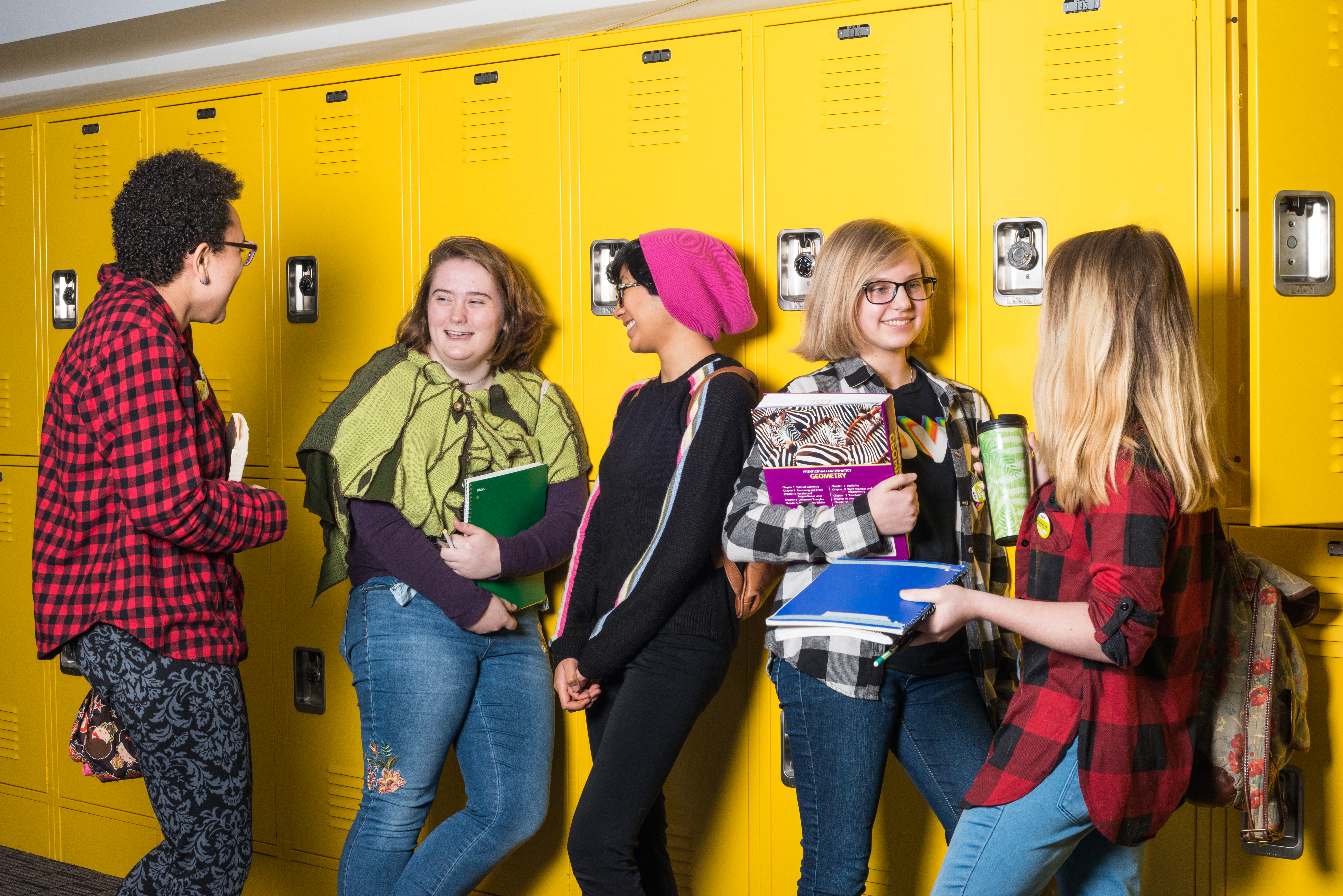 Five high school students carrying backpacks and books lean against bright yellow lockers and amicably talk to each other.