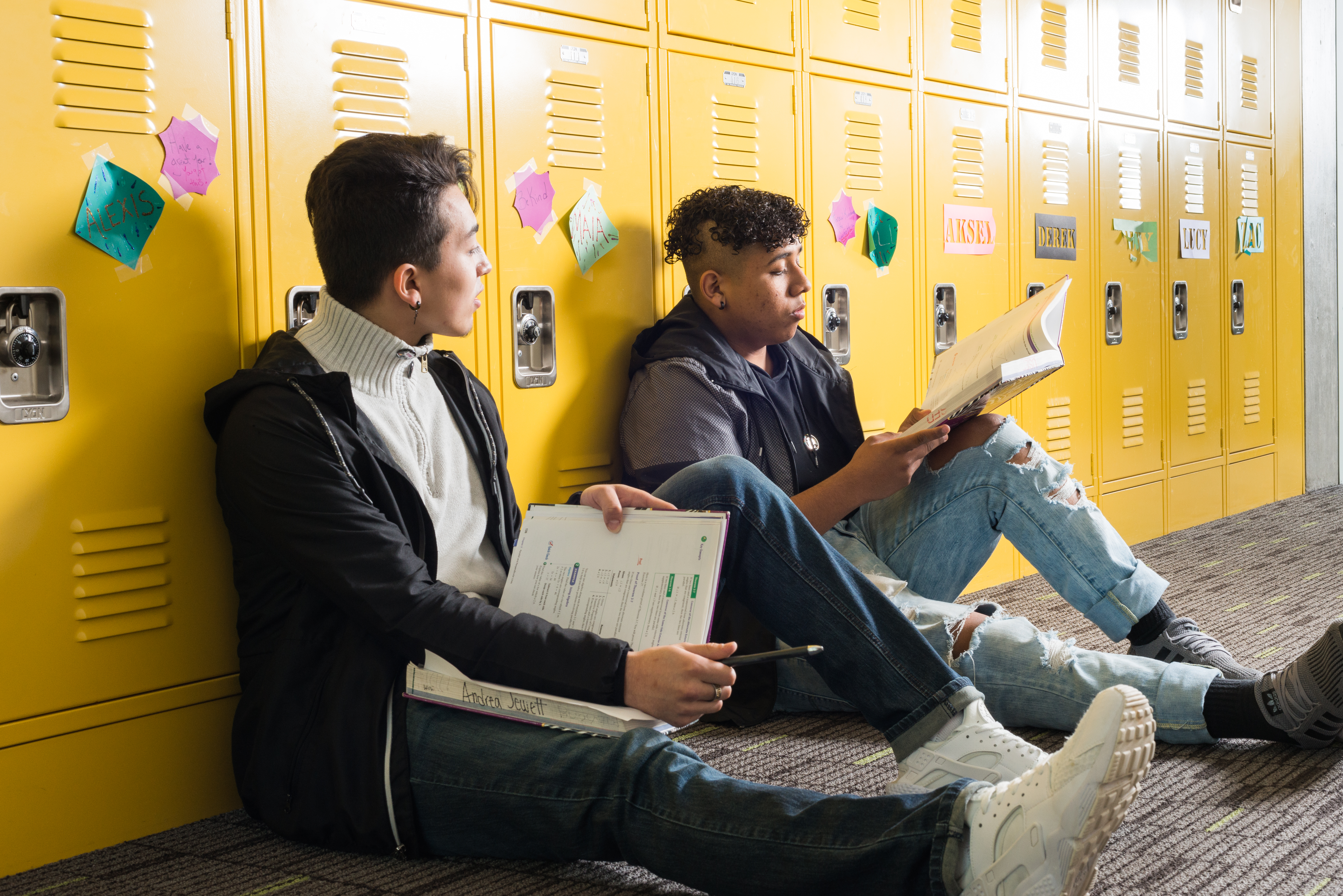 Two high school students sit with their legs stretched out against a bank of yellow lockers with post-it notes with names stuck to them. The students are studying their textbooks. One looks over at something on the other's book.