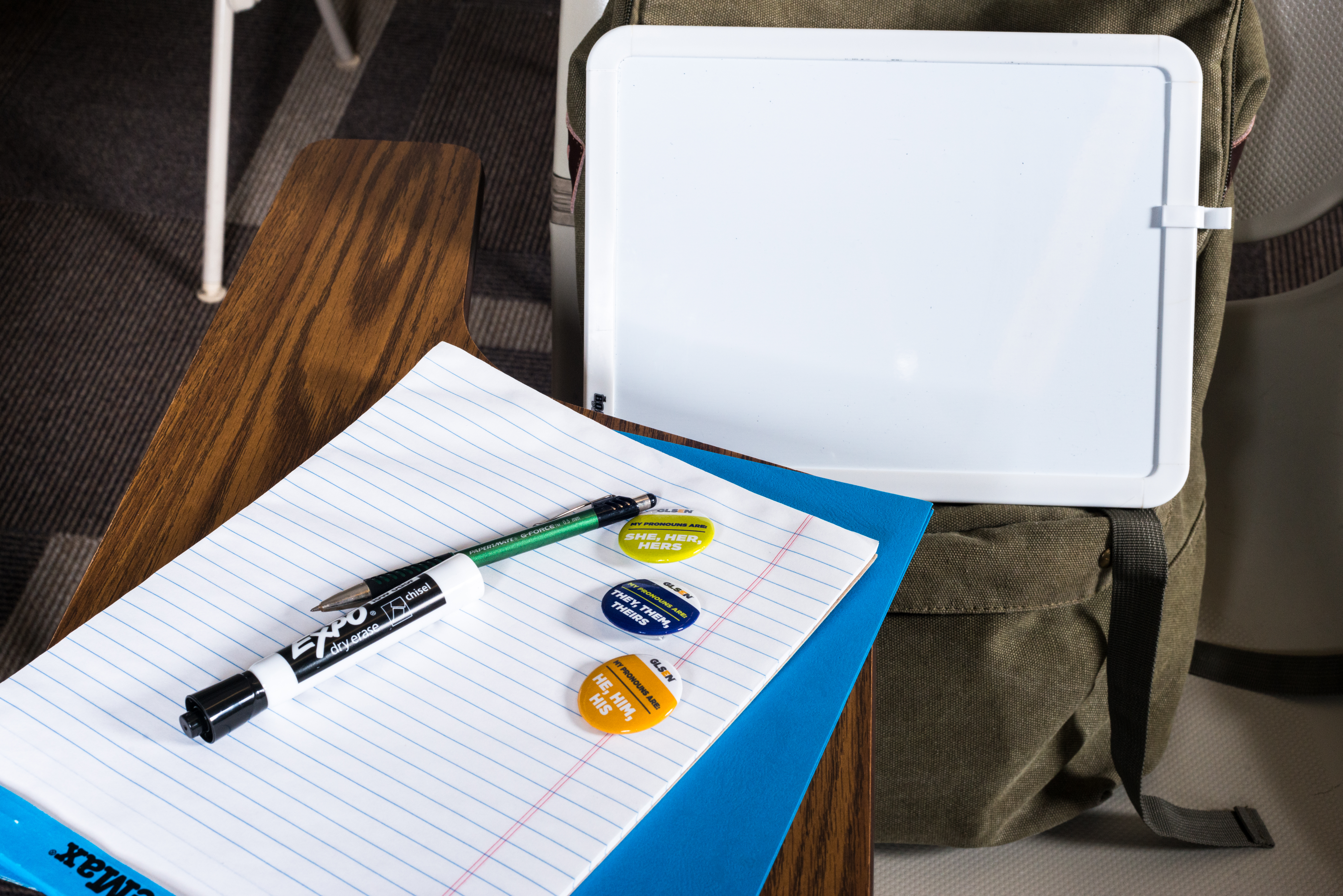 Three GLSEN pronoun buttons sit on a lined notebook on a classroom desk. From top to bottom, the buttons are lime, purple, and gold. From top to bottom, the buttons read: My pronouns are She, Her, Hers; My pronouns are they, them, theirs; My pronouns are he, him, his. Also on the desk are a folder, a dry erase marker, and a pen. On the desk chair is a backpack and a blank mini whiteboard.