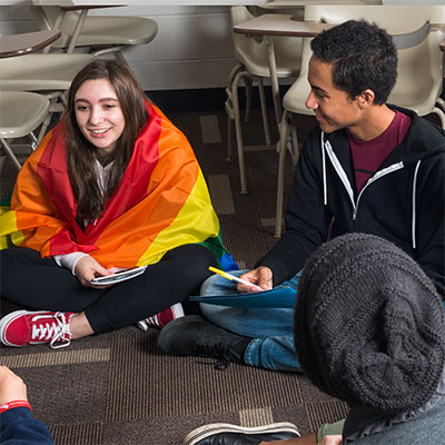 A group of students at a GSA meeting sitting on the floor. One student is wearing a rainbow flag.